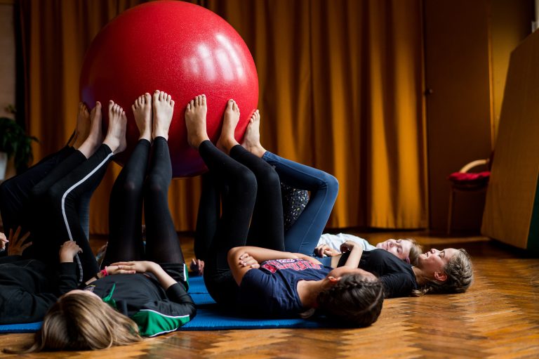 Children led on the floor in a circle holding a large ball up in the air with their feet.