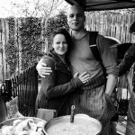 black and white image of man with an arm around a woman. In front of them a large pot of food is cooking.