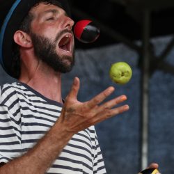 A man juggling with balls and an apple. His mouth is opened wide as if to try and catch the apple.