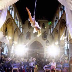 Woman acrobat hanging midair, with the backdrop of a church behind her.